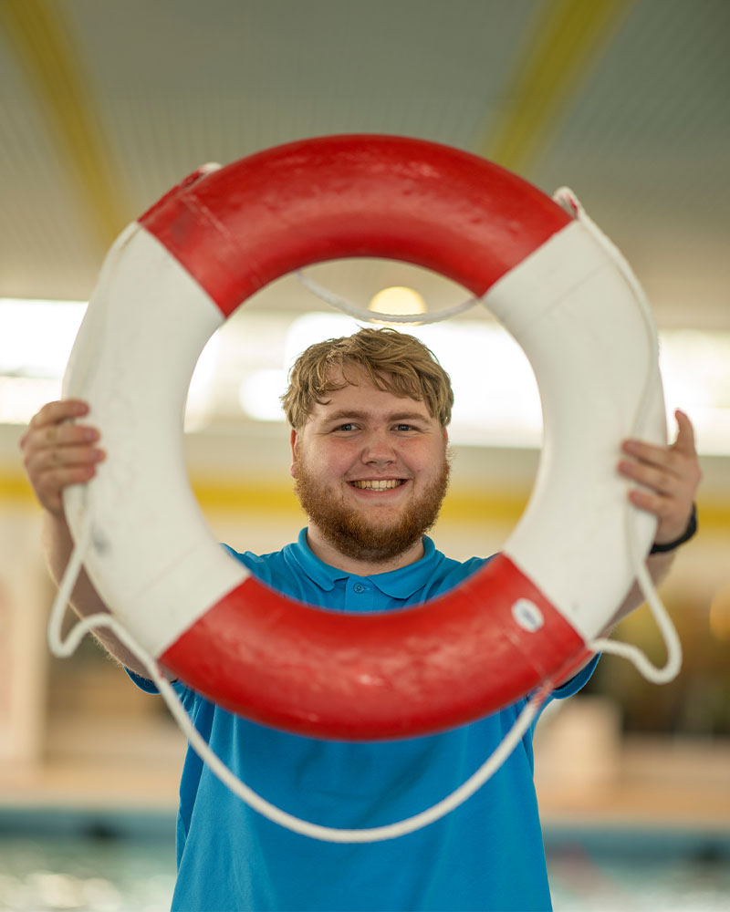 Der Mitarbeiter des Freizeitbads Aquarell, Robin, hält lächelnd einen Rettungsring in die Kamera